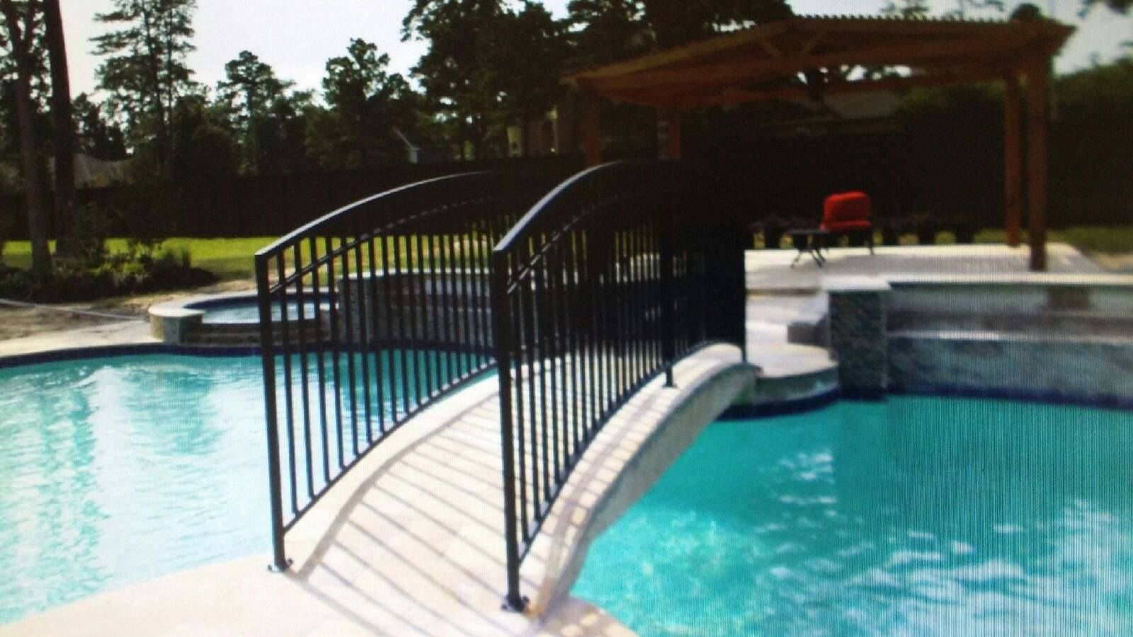 A black metal arched footbridge spans across a turquoise swimming pool in a backyard with a wooden pergola.