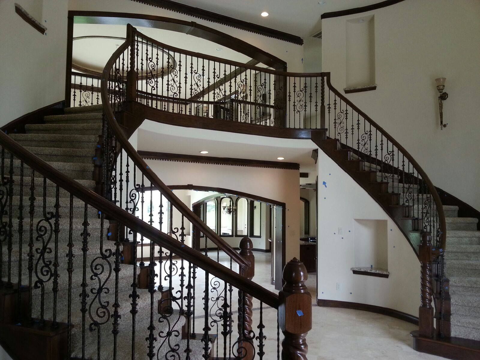 A grand, curved wooden staircase with ornate iron railings connects to an upper landing in a light-toned home foyer.
