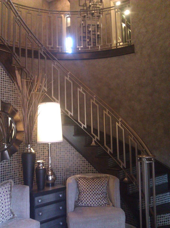 A curved staircase with geometric metal railings stands behind a small table with a lamp, vase, and two armchairs.