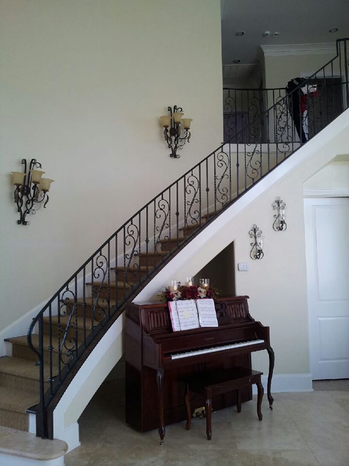 A piano sits under a staircase with a black iron railing, next to a hallway with wall sconces and hanging wall decor.