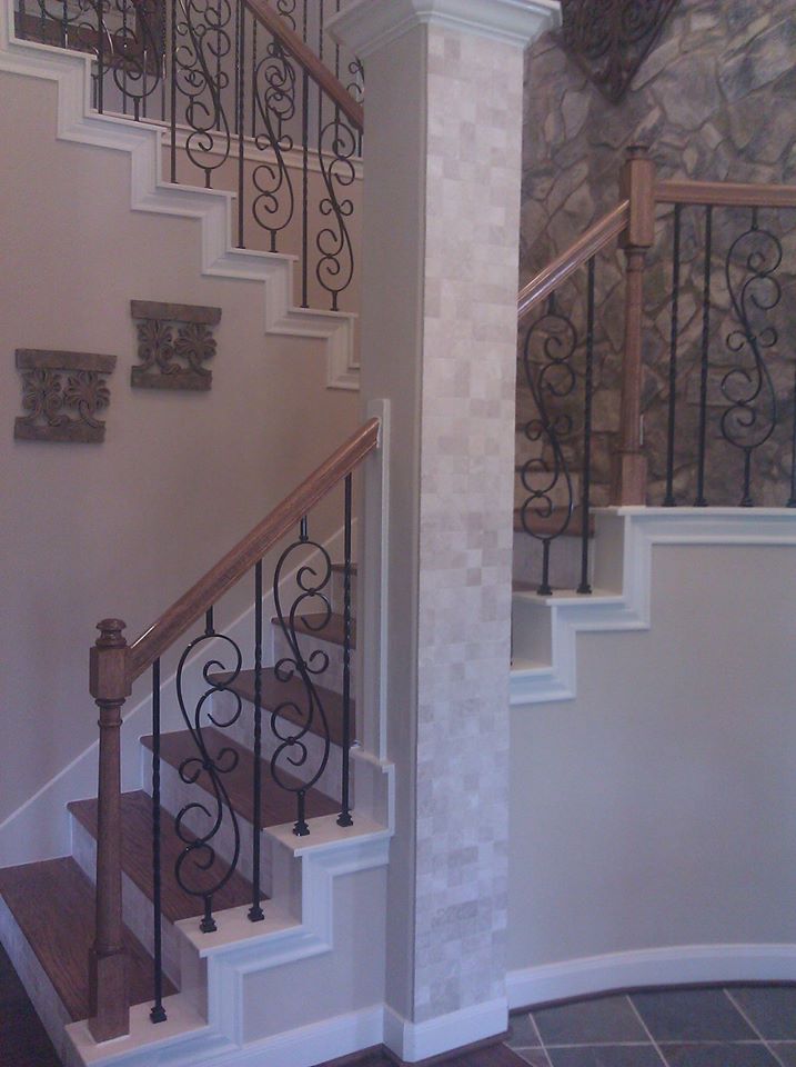 A split staircase featuring wooden handrails, dark iron scroll balusters, tan walls, and a stone-tiled pillar.