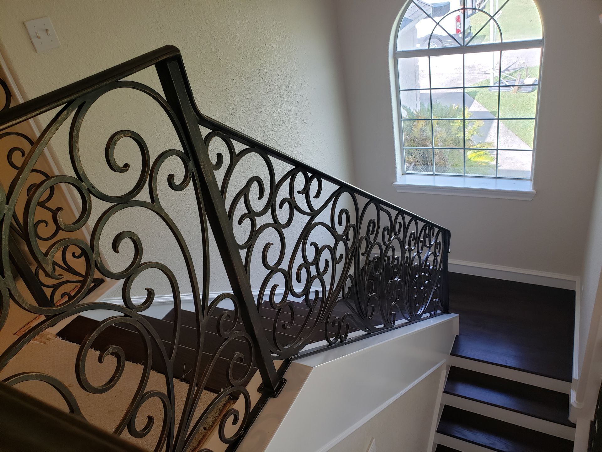 A staircase with dark wooden treads and white walls features an ornate black wrought-iron railing beneath a arched window.