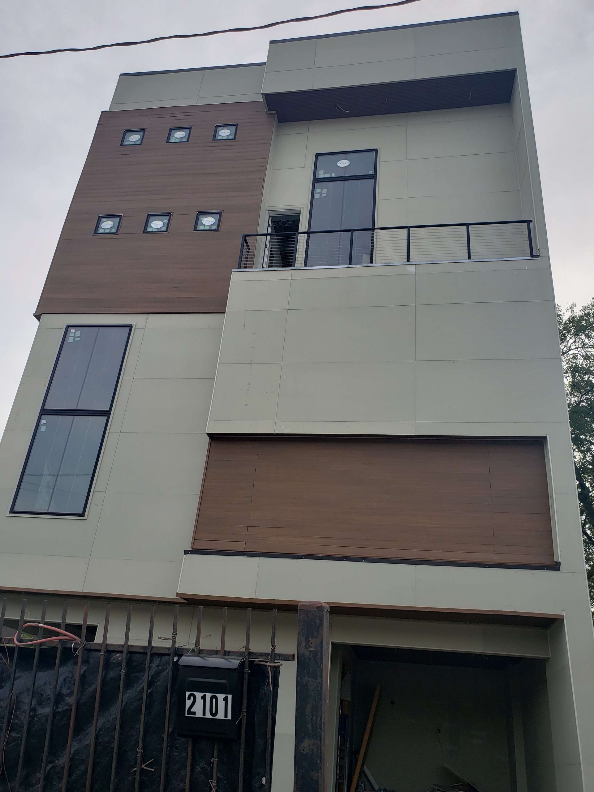 A three-story modern building with beige stucco walls, dark wood accents, a balcony, and a fenced-in lower entrance.