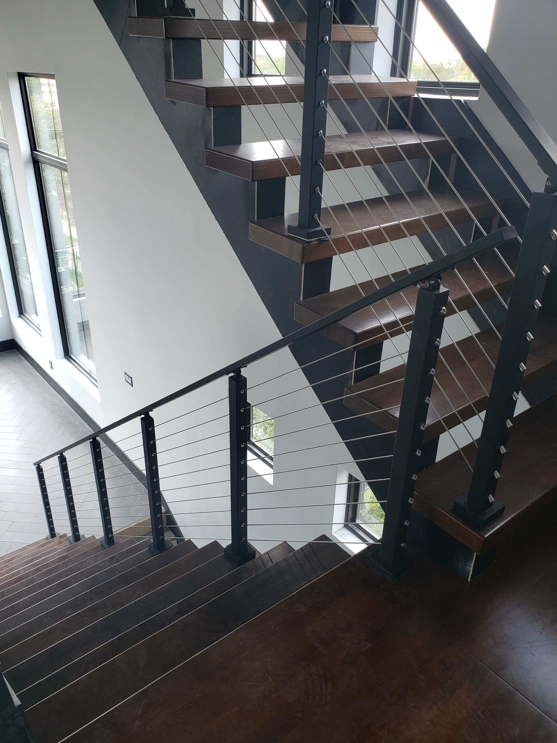A high-angle view of a wooden staircase with a modern black metal frame and cable railing, viewed from the upper landing.