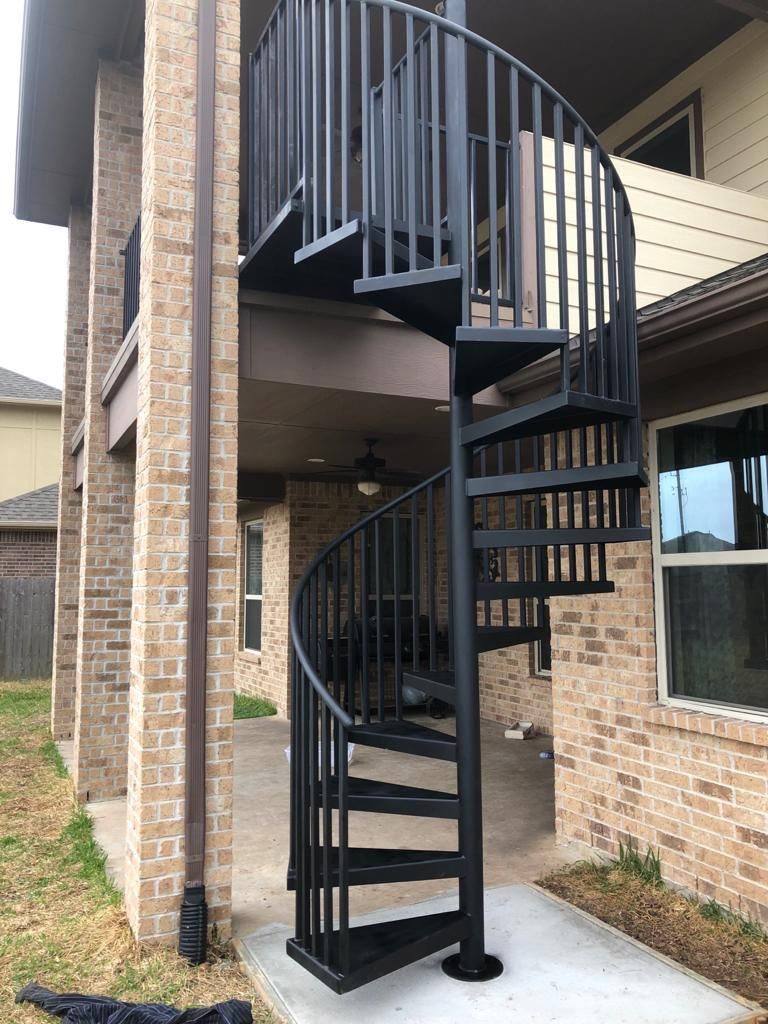 A black metal spiral staircase installed on a concrete patio next to a brick house.