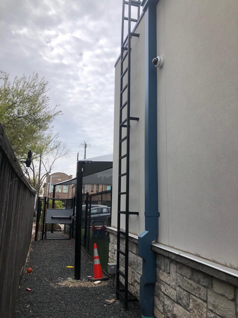 A black metal ladder is mounted to the side of a light-colored stucco and stone building, beside a blue downspout.