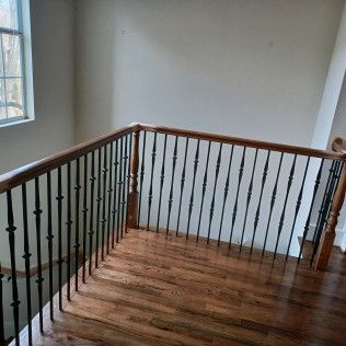A wooden banister with black metal spindles and hardwood flooring in an indoor hallway next to a window.