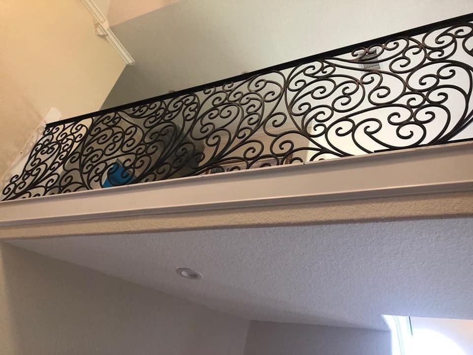 An interior view of a black metal scrollwork railing along a balcony overlooking a room with white walls and ceiling.