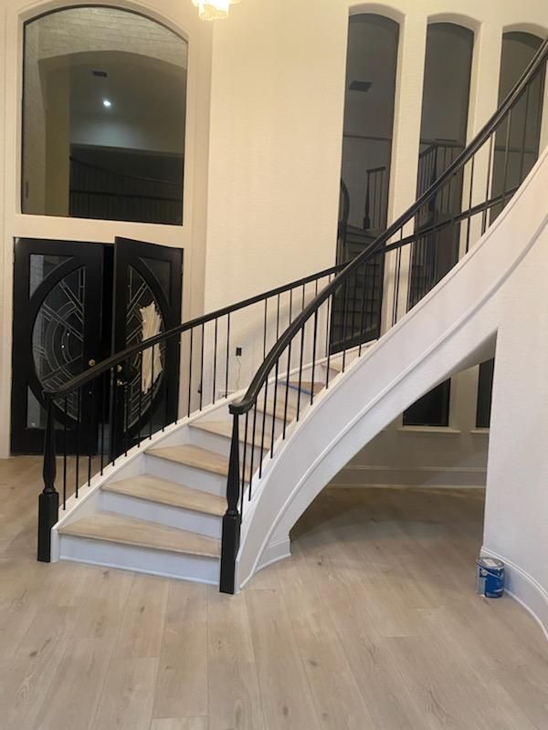 A curved staircase with wooden treads and black iron railings in a bright, modern foyer with double doors.