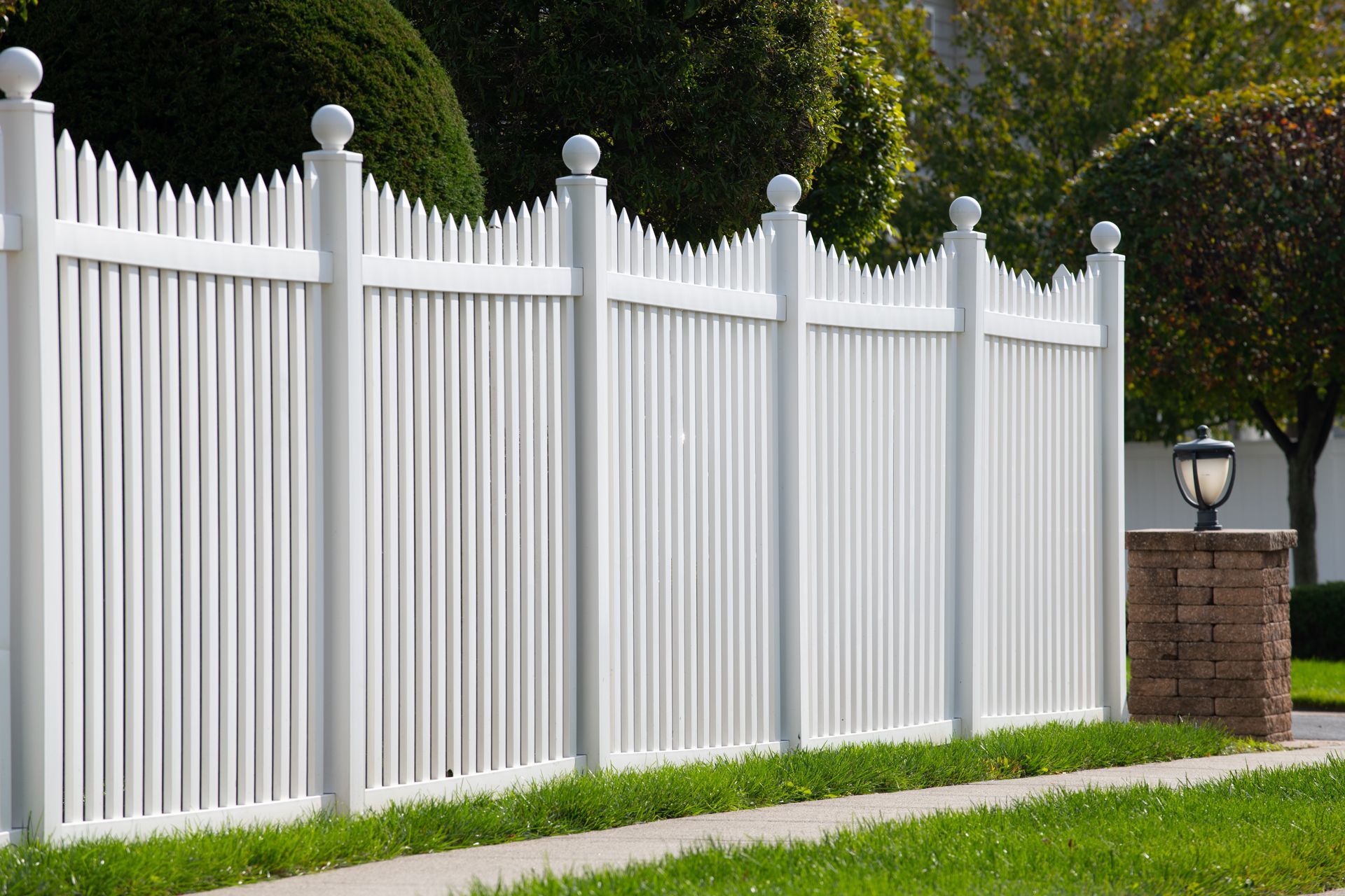 A white picket fence with decorative ball-topped posts runs along a green lawn and sidewalk on a sunny day.