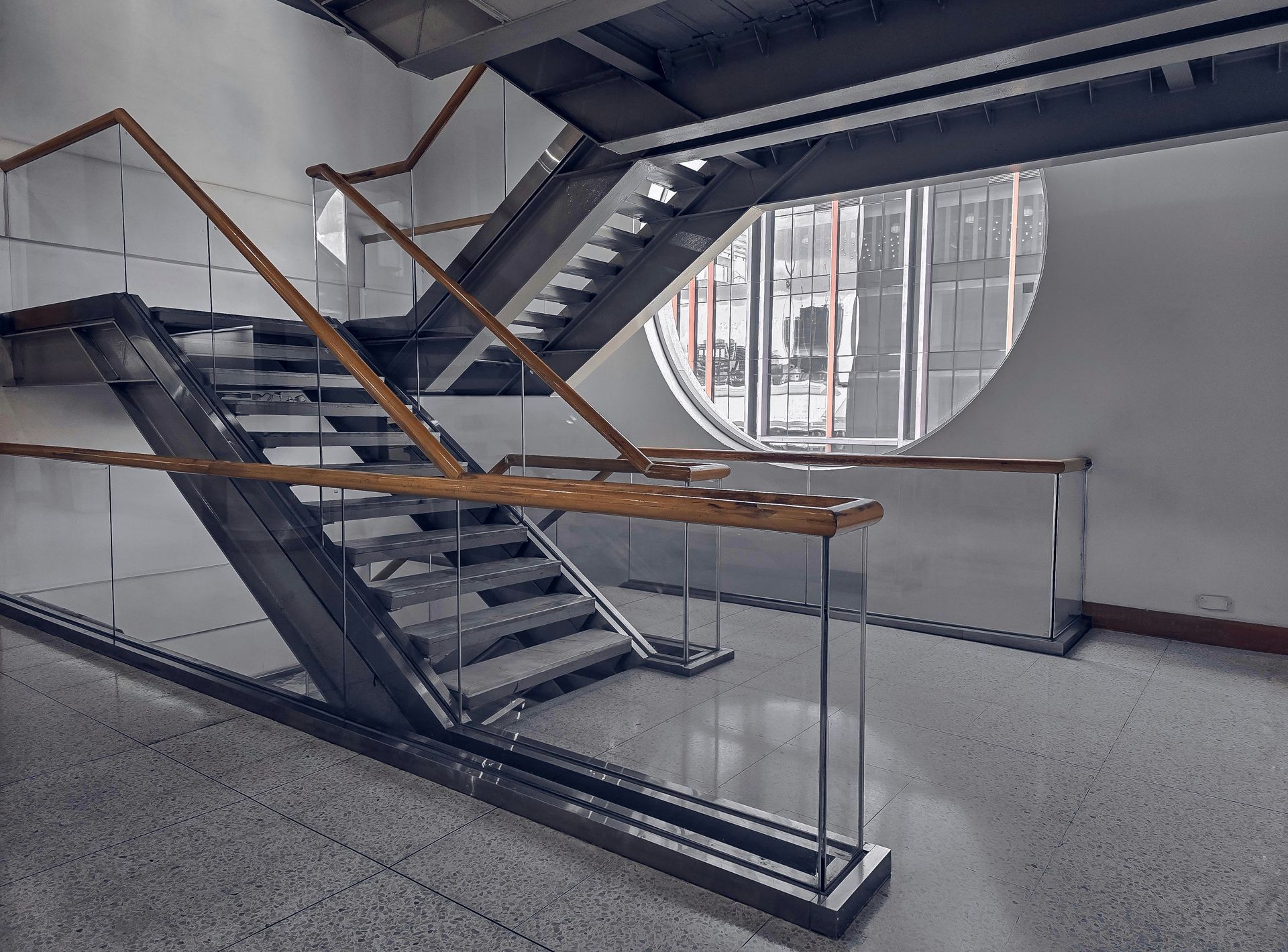 A staircase with metal frame, wood handrails, and glass panels leads to an upper level, beside a round window.