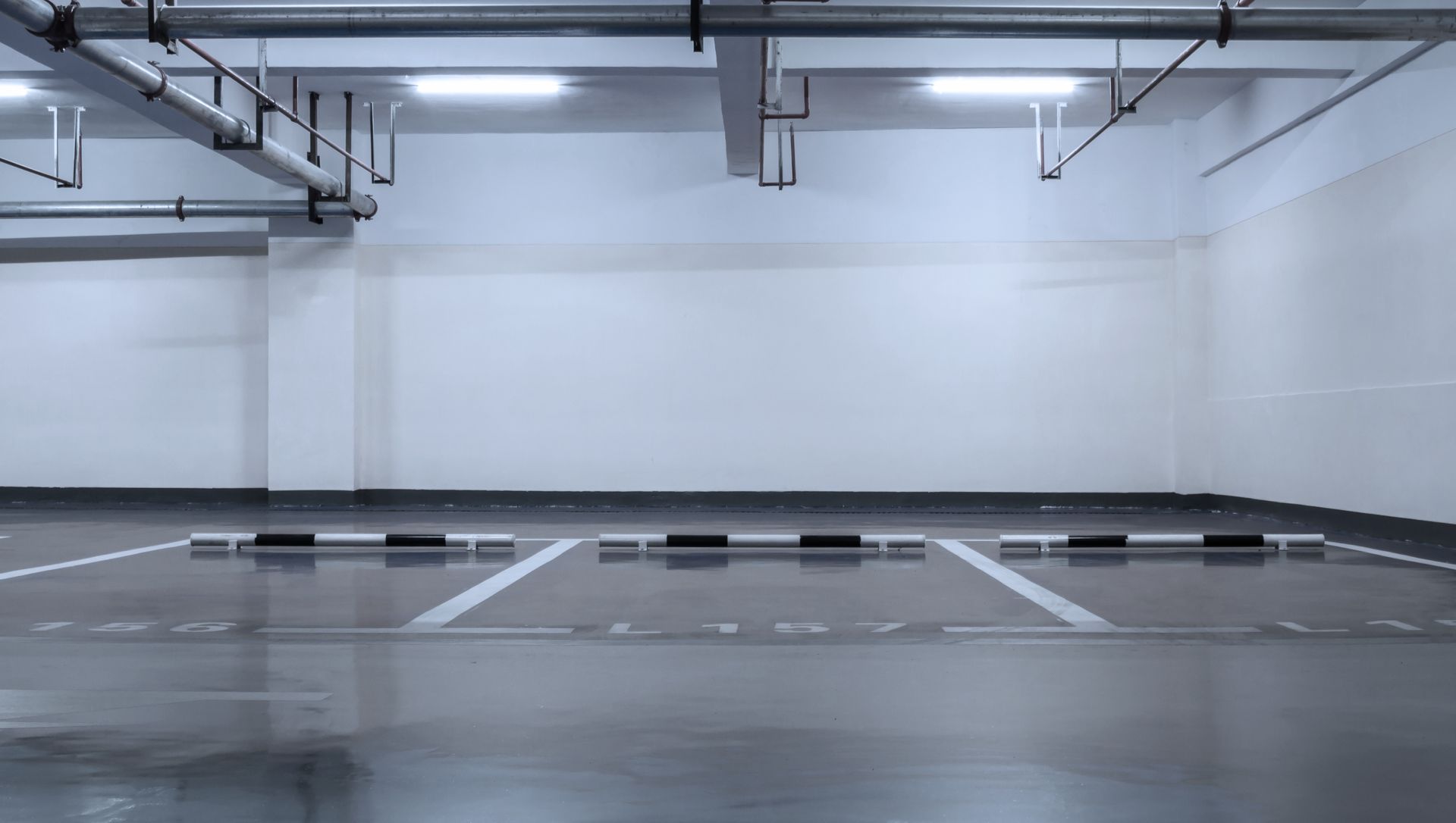Empty, well-lit parking garage. White walls, reflective gray floor, overhead pipes, and parking space markings.
