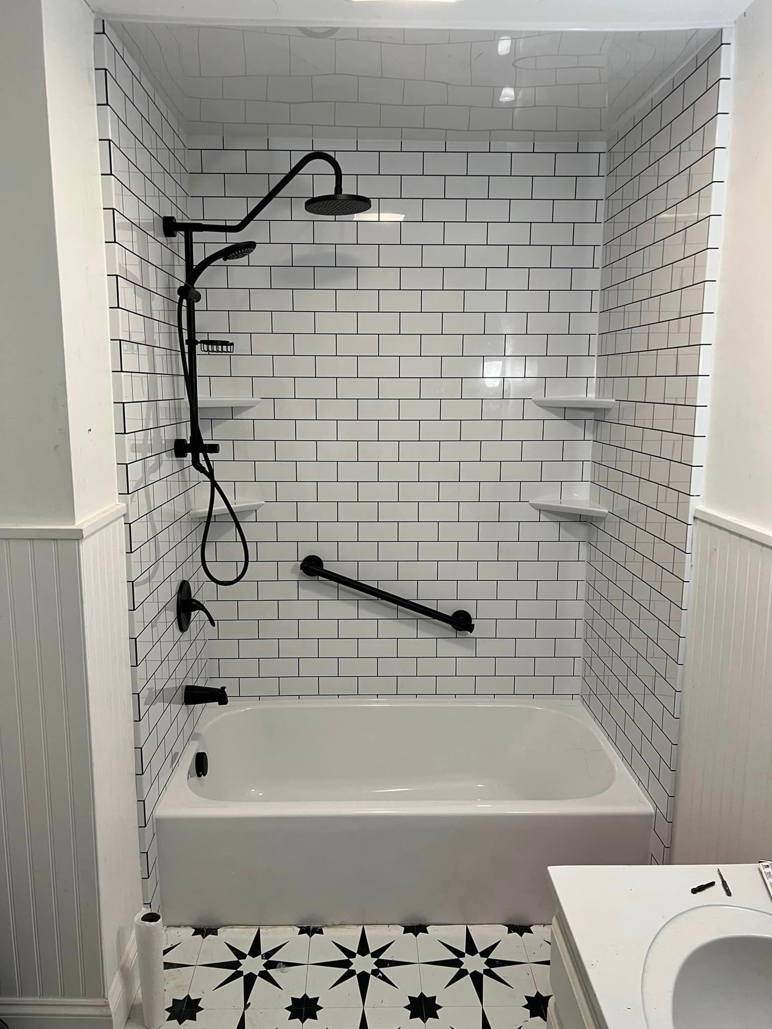 A modern bathroom with white subway tile and black accents, including a shower head, grab bar, and decorative floor tiles.