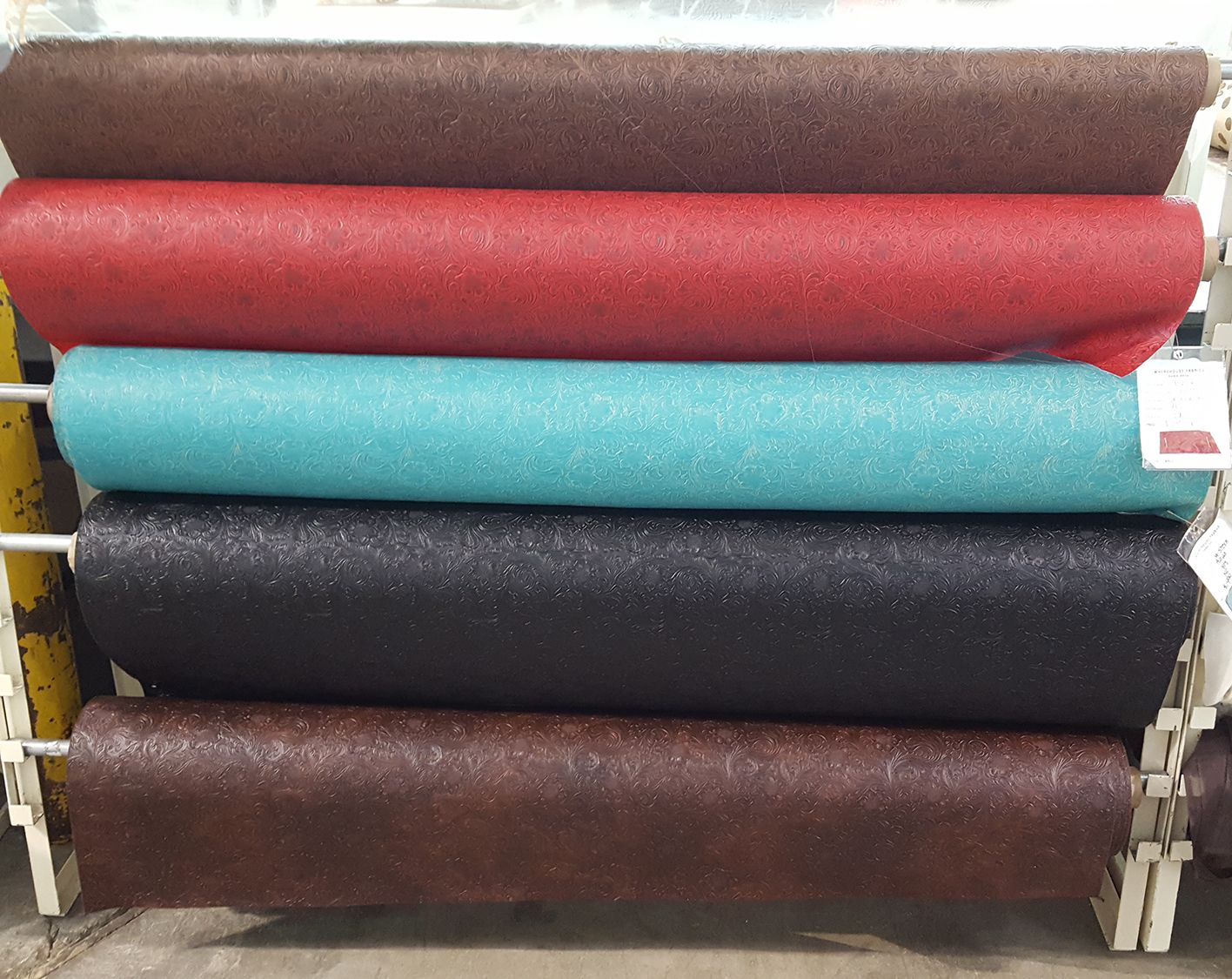 Shelving unit holding five rolls of textured fabric in various colors: brown, red, teal, black, and dark brown.