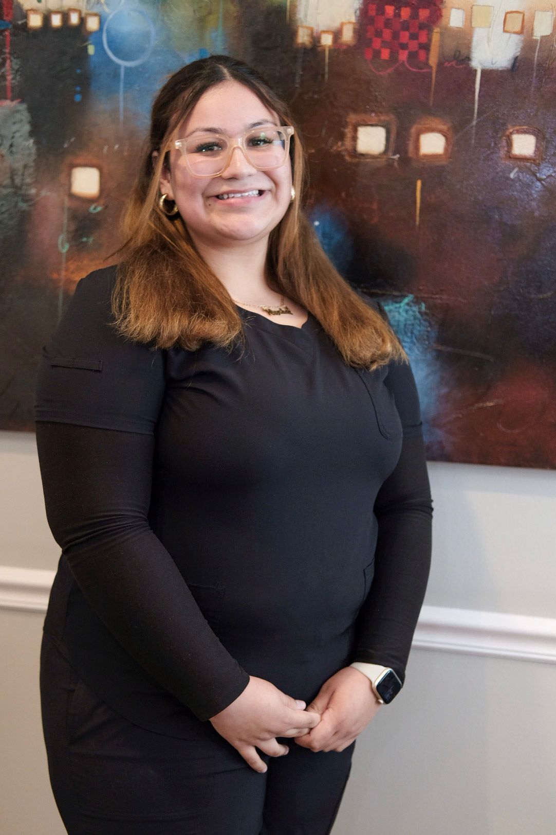 A woman with long curly hair is wearing a black scrub top and smiling.