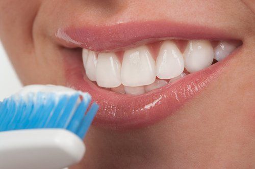 Person smiling with bright white teeth, applying toothpaste from a blue toothbrush.