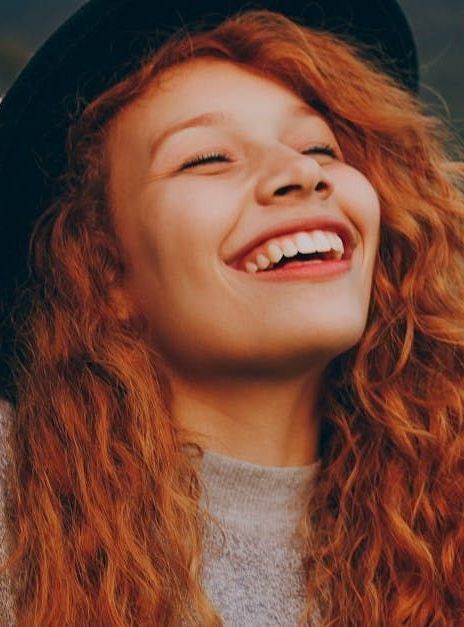 A woman with red hair is wearing a black hat and smiling.