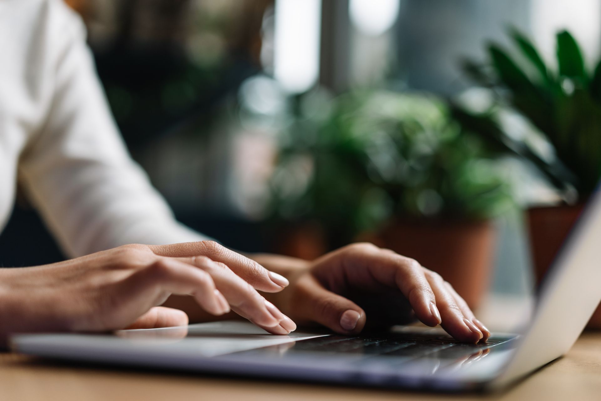 Person's hands typing on a laptop keyboard, brown table, blurred green plants in background.