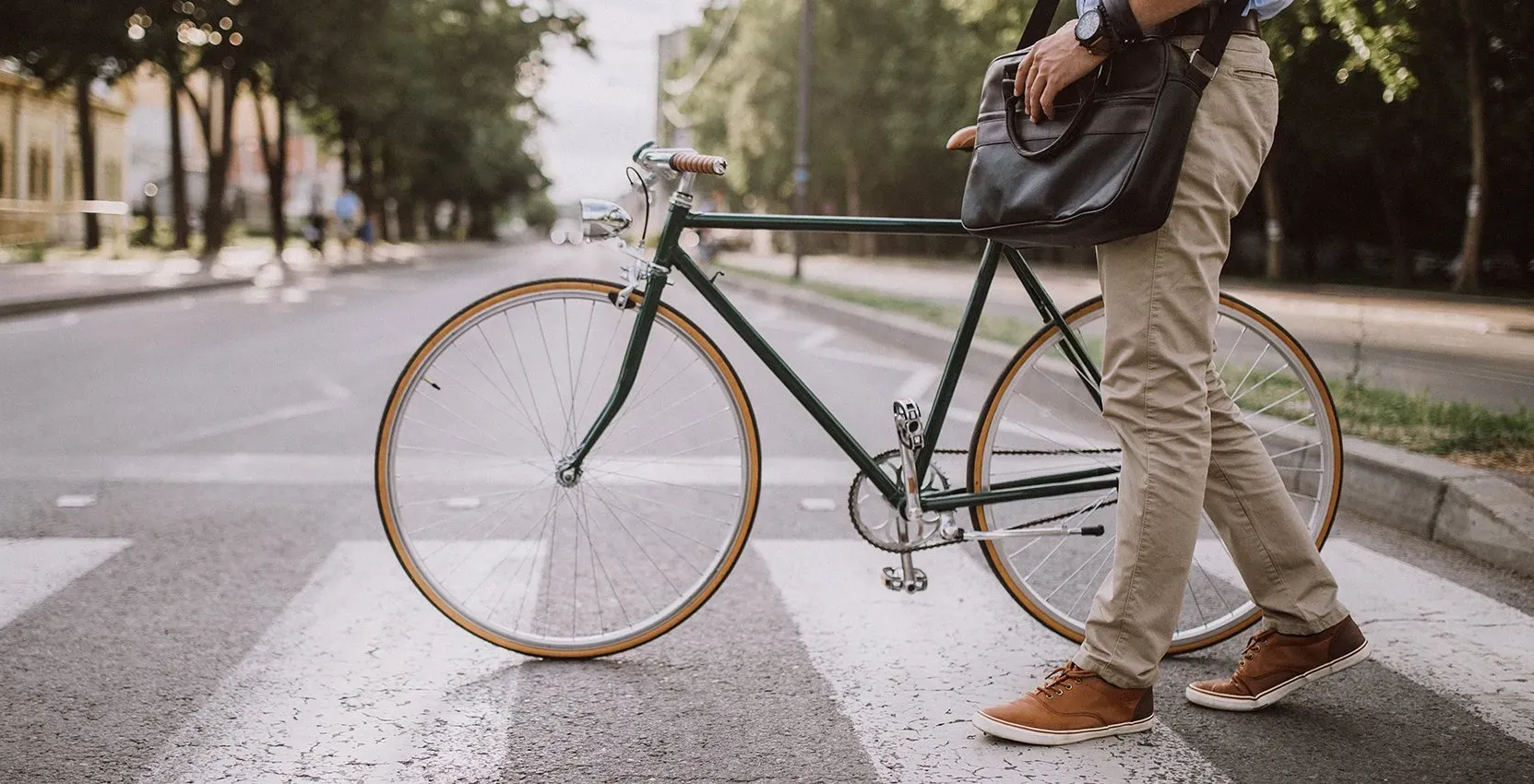 Man walking across crosswalk with bicycle, wearing tan pants and carrying a bag.