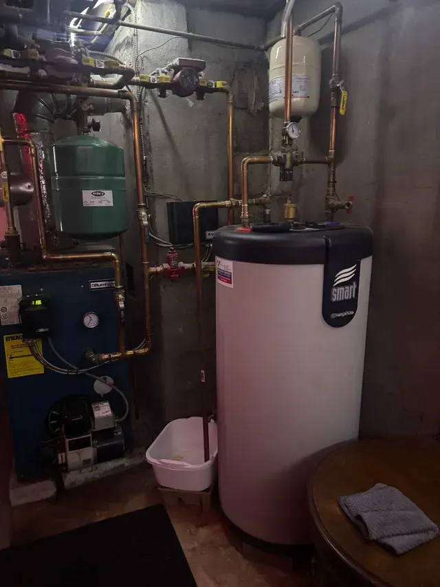 Boiler room with a water heater, copper pipes, and a white bucket. Dark, concrete walls and floor.