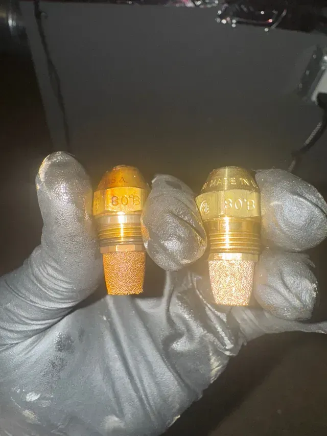 Two brass oil burner nozzles held in gloved hands, showing a porous filter.
