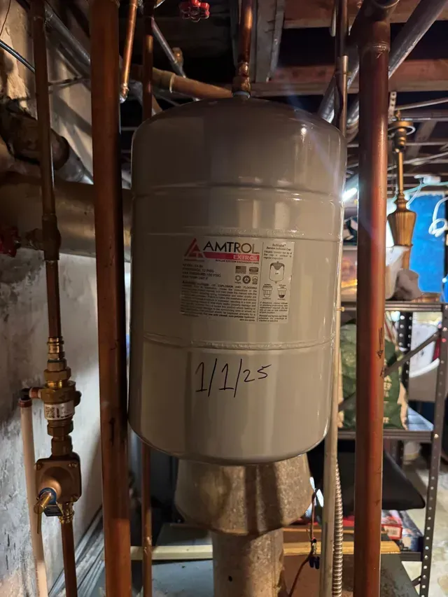 A vertical, gray thermal expansion tank for a heating system in a basement, with copper pipes.
