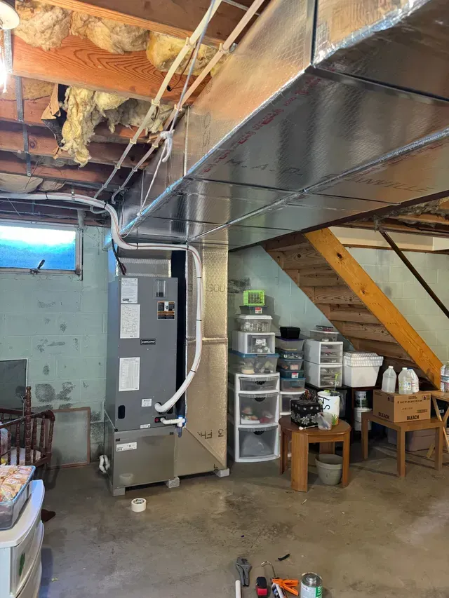 HVAC unit in a basement. Ductwork overhead. Stairs and storage on the right, partially visible.