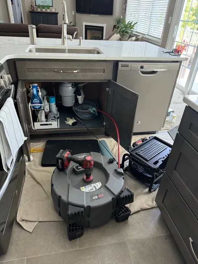Plumber unclogging kitchen sink drain with a snake machine. Open cabinet, gray cabinets, and a dishwasher are visible.