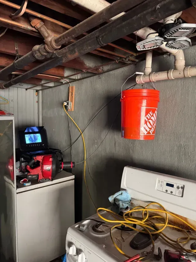 Basement scene with pipes, a washing machine, and an orange bucket hanging. Plumbing equipment on white cabinet.
