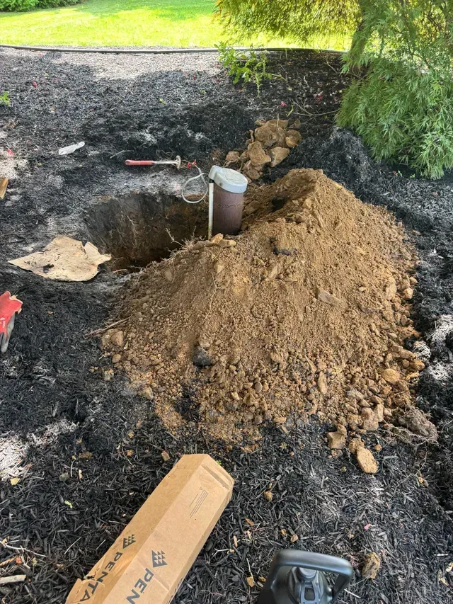 Hole dug in a garden bed with a well or pipe exposed. A pile of dirt and tools sit nearby.