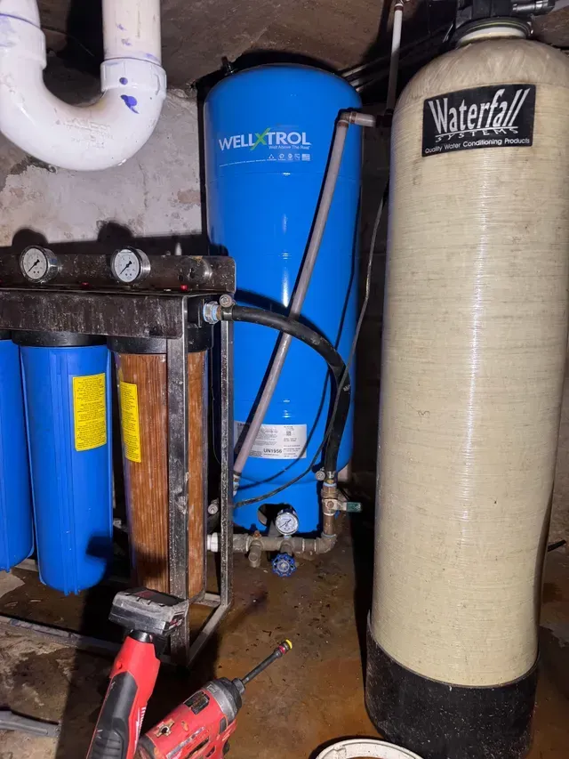 Plumbing equipment in a basement, including a blue Well-Trol pressure tank, water filter, and a waterfall water softener.