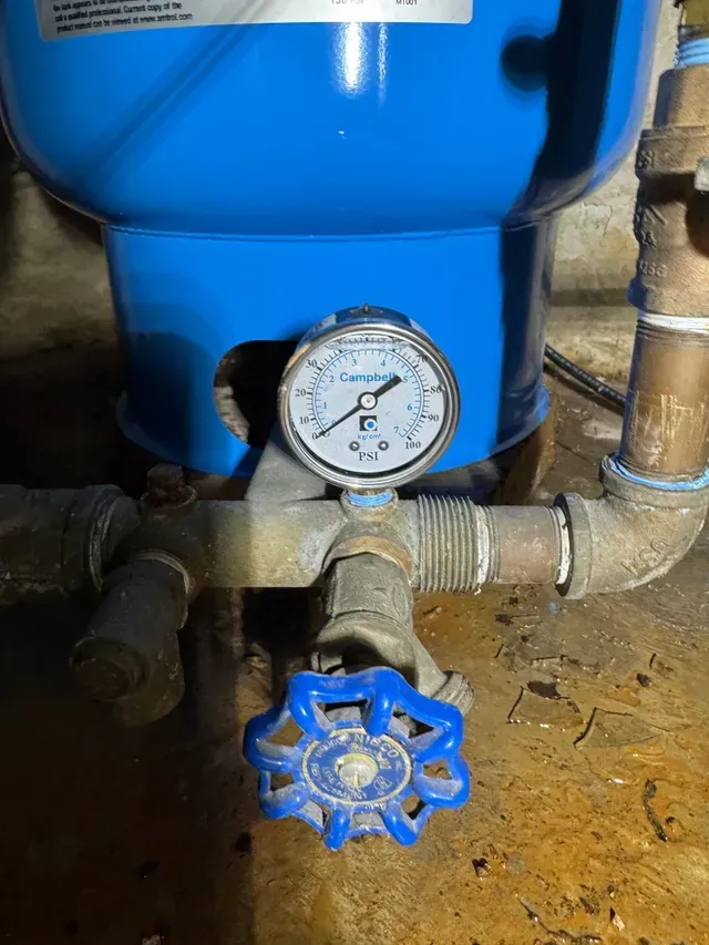 Blue water pressure tank with gauge, valve, and pipes in a basement.