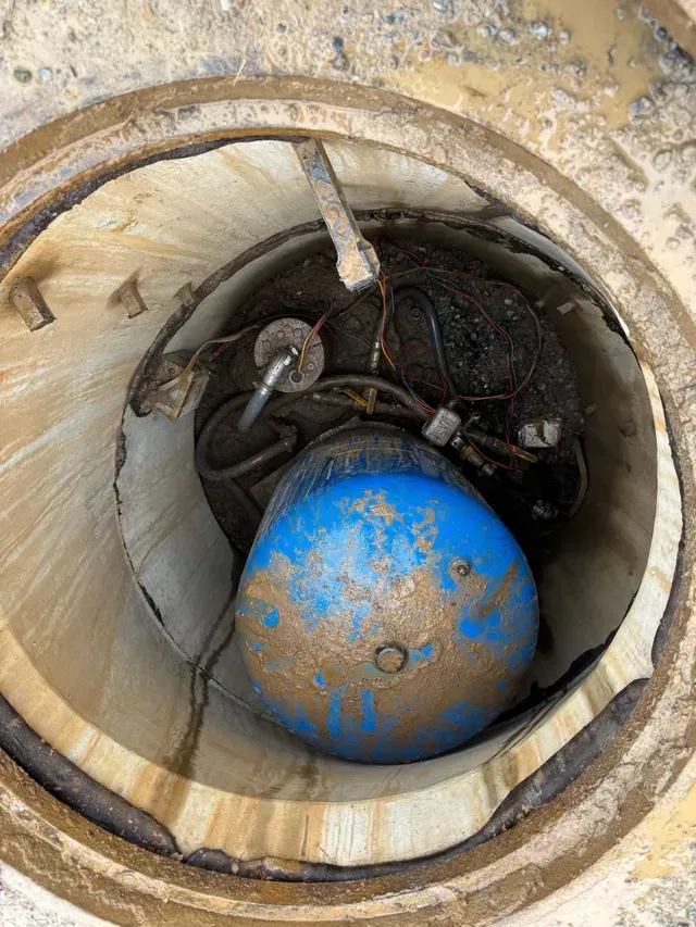 Open manhole revealing a blue, round object, possibly a pump, covered in grime.