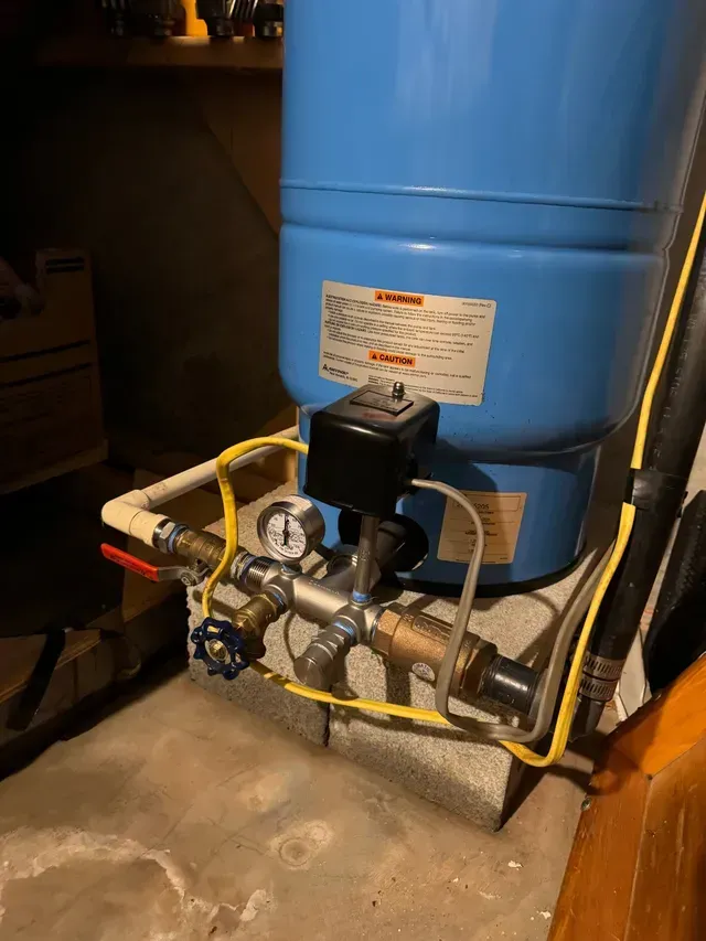 Blue water pressure tank with gauges, valves, and electrical components on a concrete block in a basement.