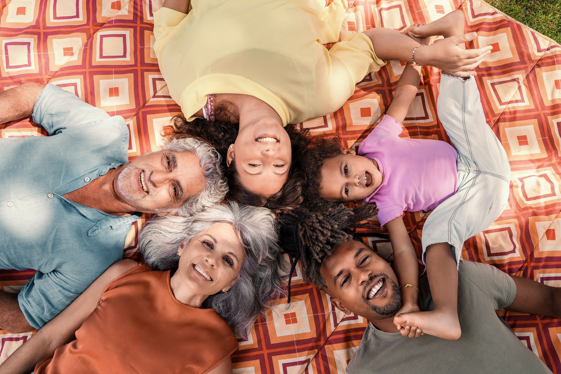 Family laying on a red and orange blanket outdoors; smiling faces, arms and legs touching.
