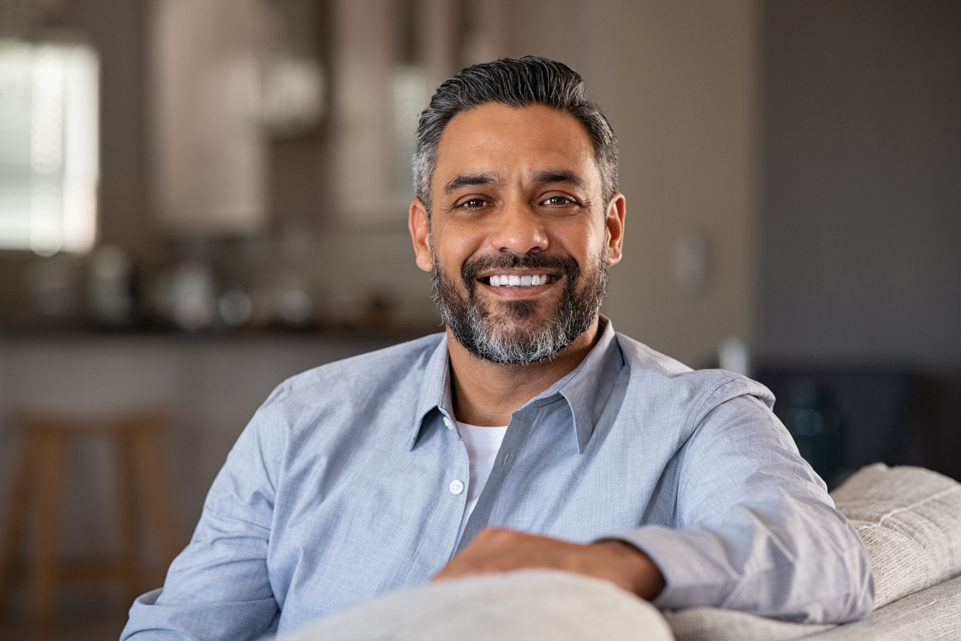 Middle-aged man with graying beard, smiling. Sitting on a couch in a home.