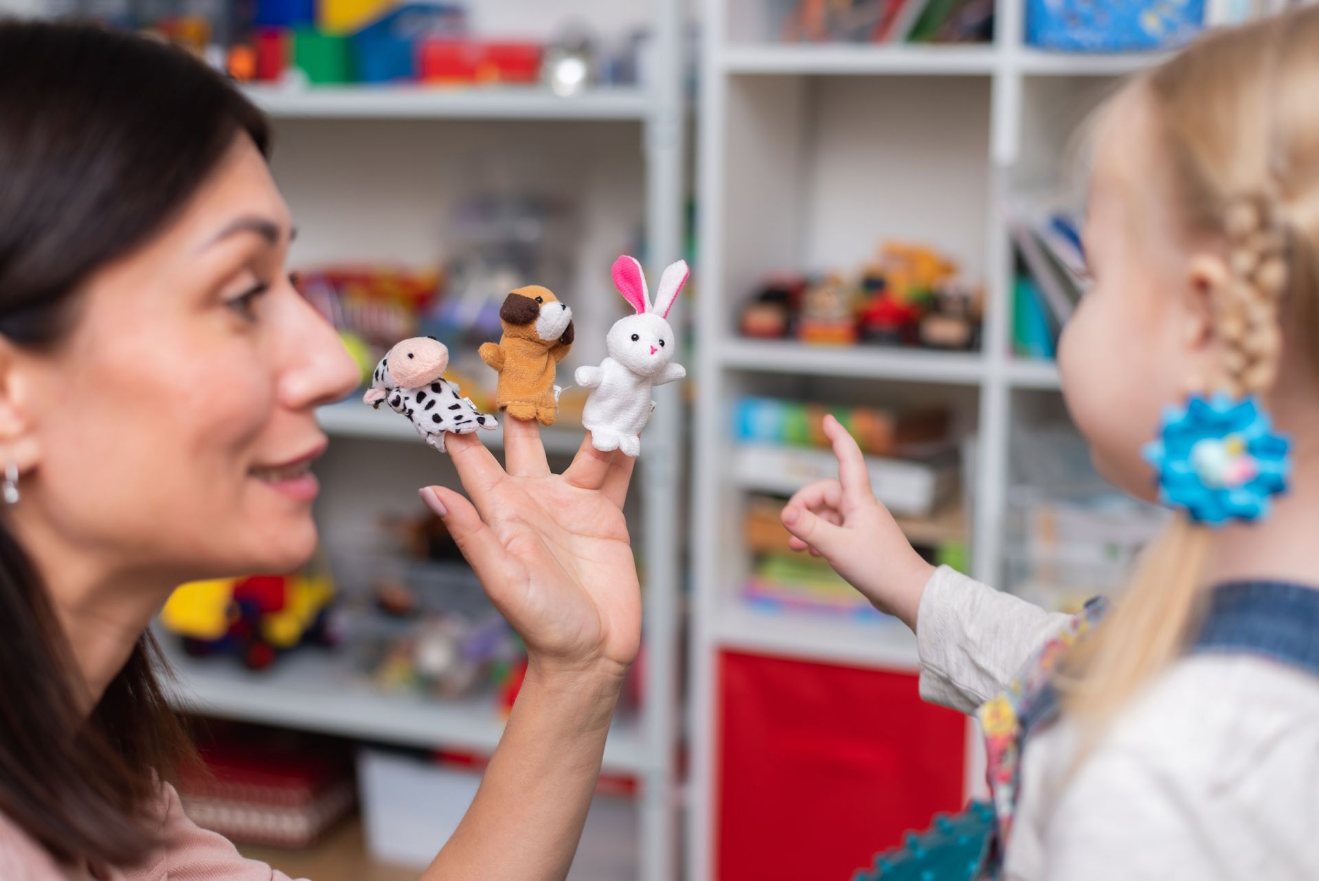 Woman using finger puppets, talking to a child with a finger pointed up; toys in the background.