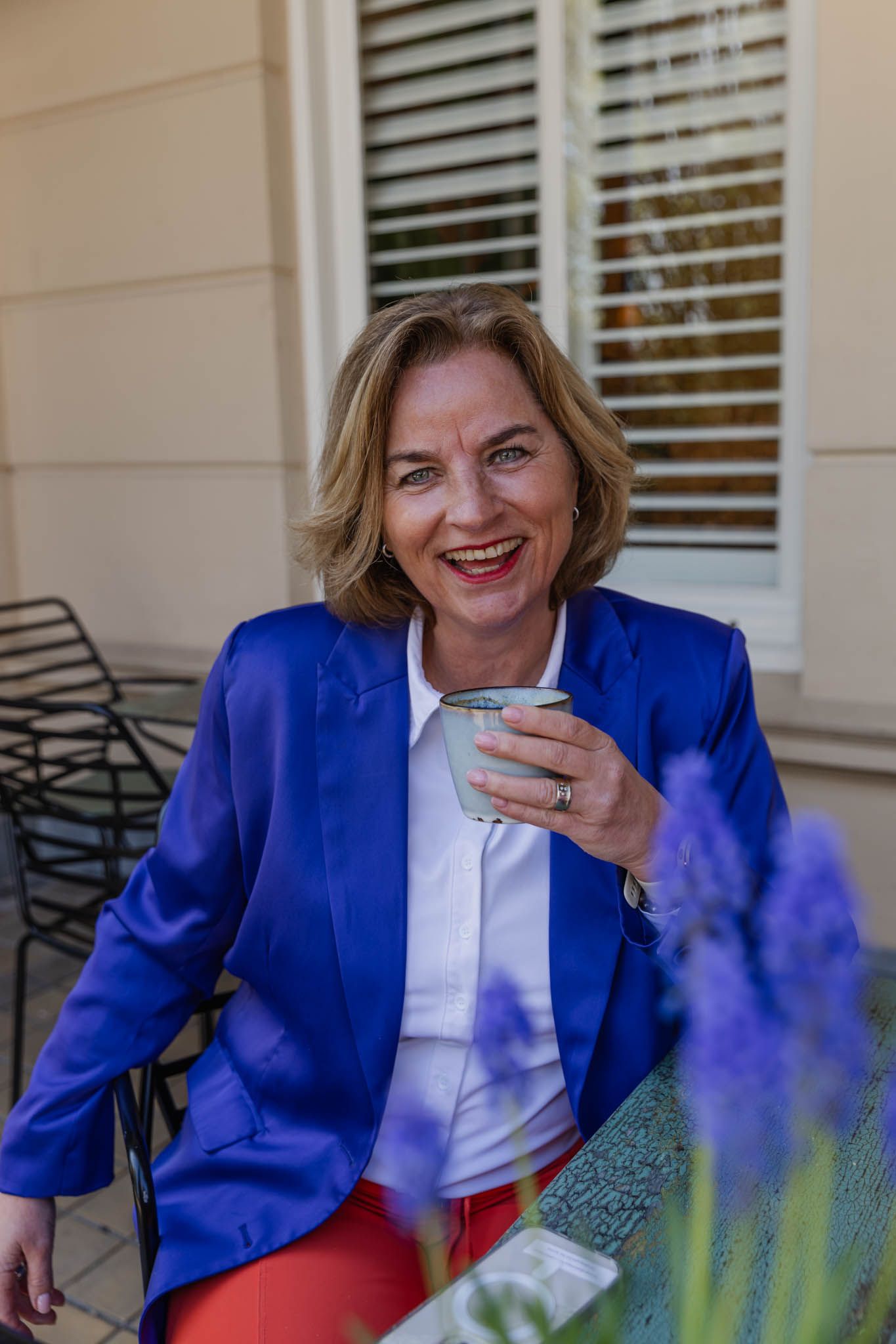 Trouwambtenaar in prive houding afgebeeld. Kopje koffie in de hand, blauwe blazer, witte blouse en oranje broek aan. Zittend aan een tafeltje.