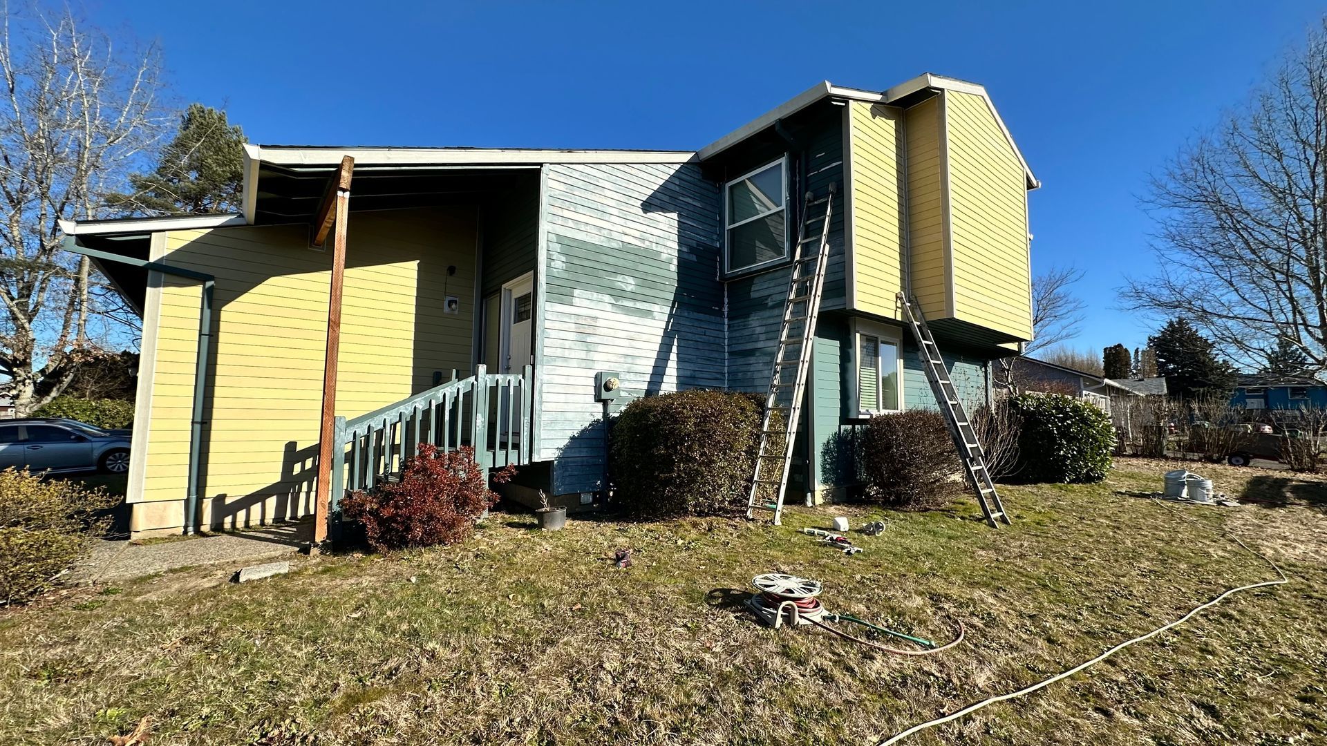 A yellow and blue house with a ladder in front of it is being painted.