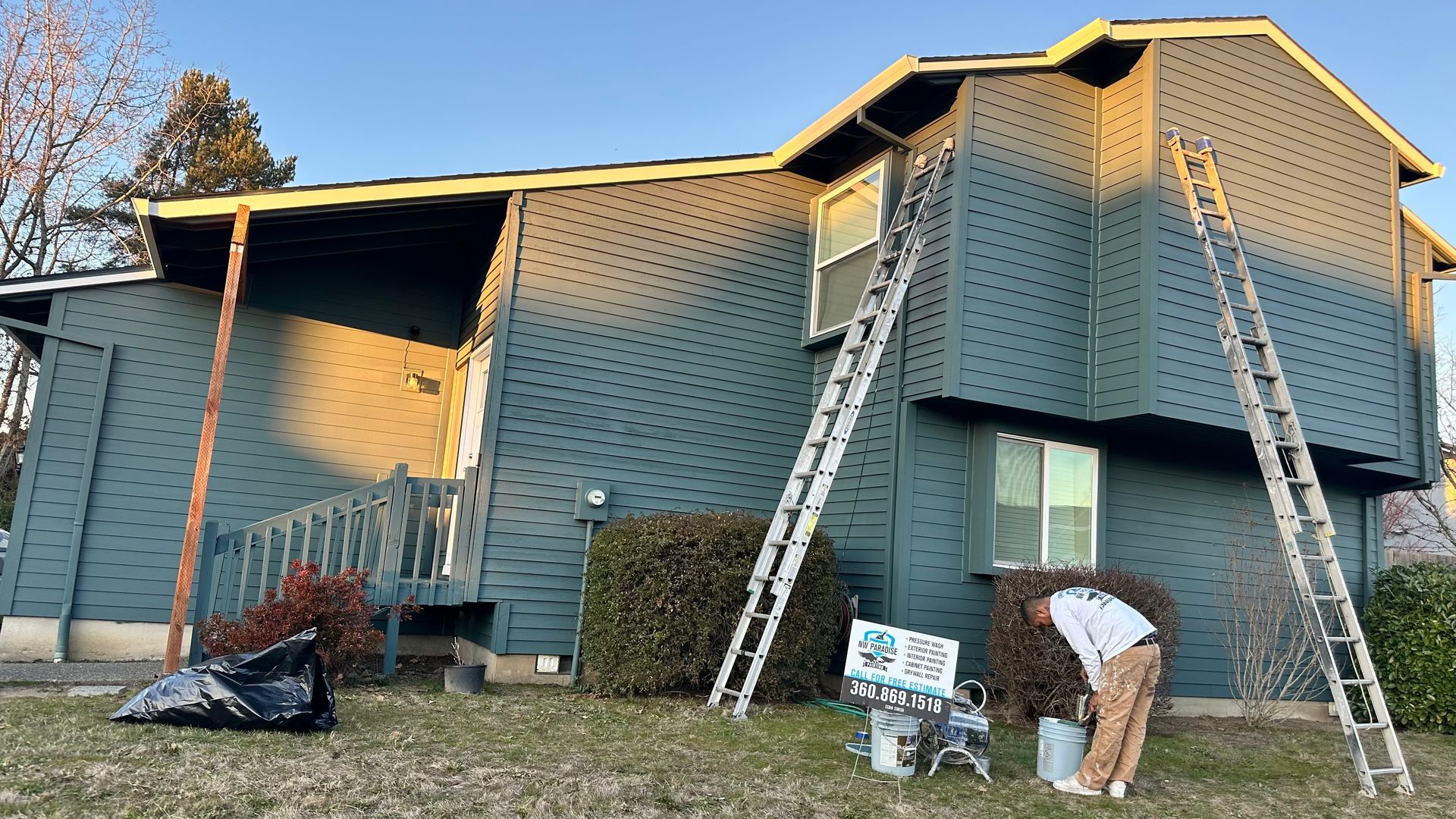 A man is painting the side of a house with ladders.