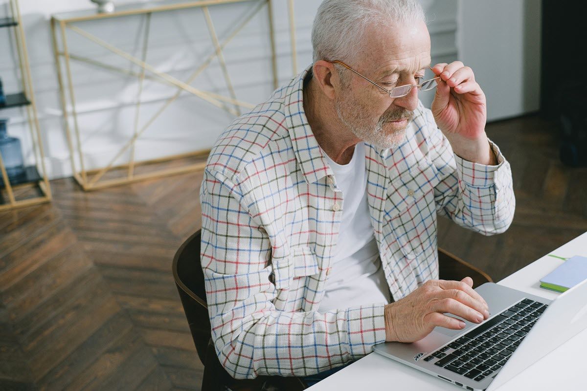 Older man in glasses, using a laptop at a desk, adjusting glasses.