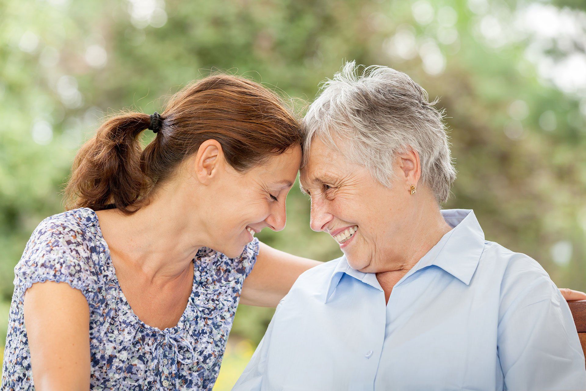 Una donna con i capelli castani e una donna anziana con i capelli grigi si toccano la fronte e sorridono, all'aperto.
