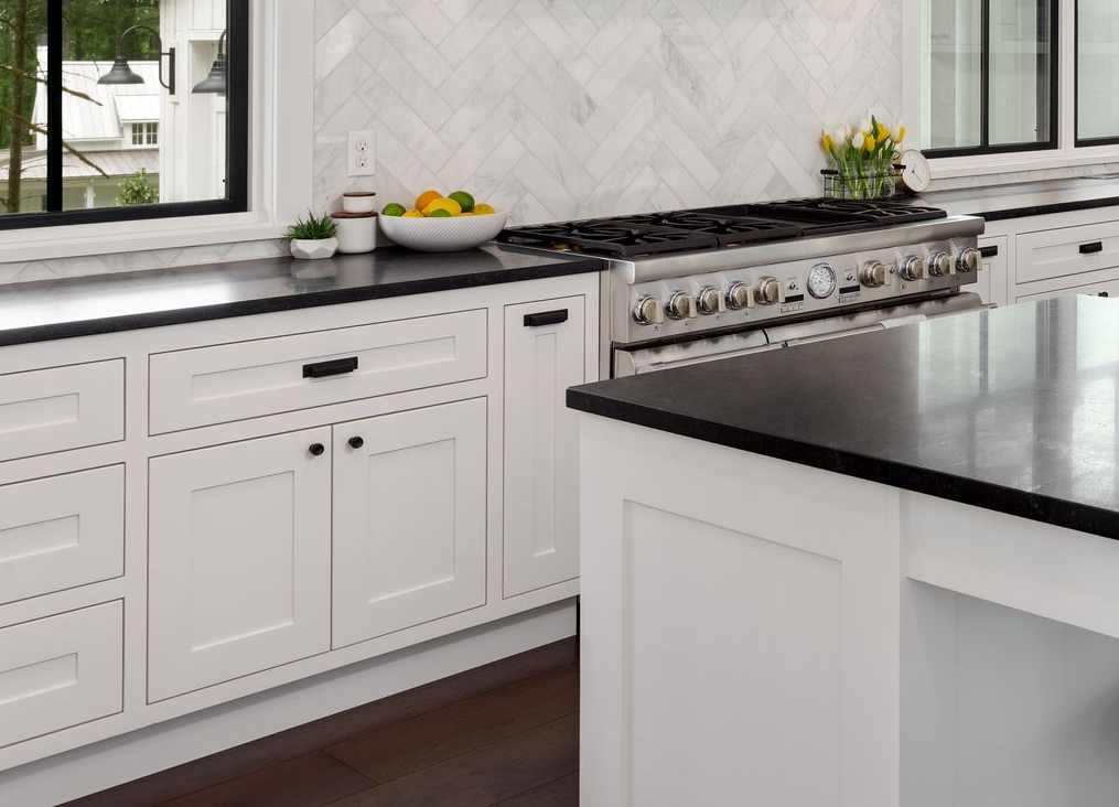Modern white kitchen with black countertop, stainless steel stove, and fruit bowl by the window