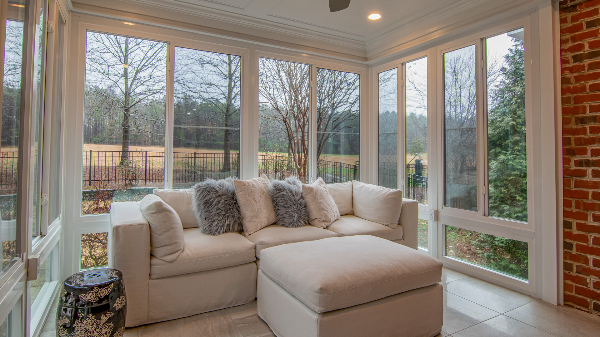 Bright sunroom with beige sectional sofa, large windows, tile floor, and brick accents overlooking trees