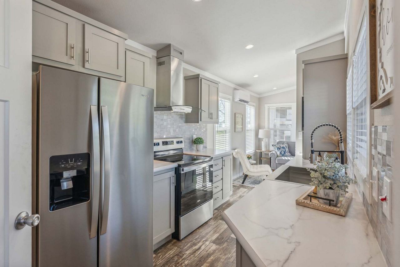 Bright white galley kitchen with stainless fridge, stove, and narrow countertop opening to a dining area.