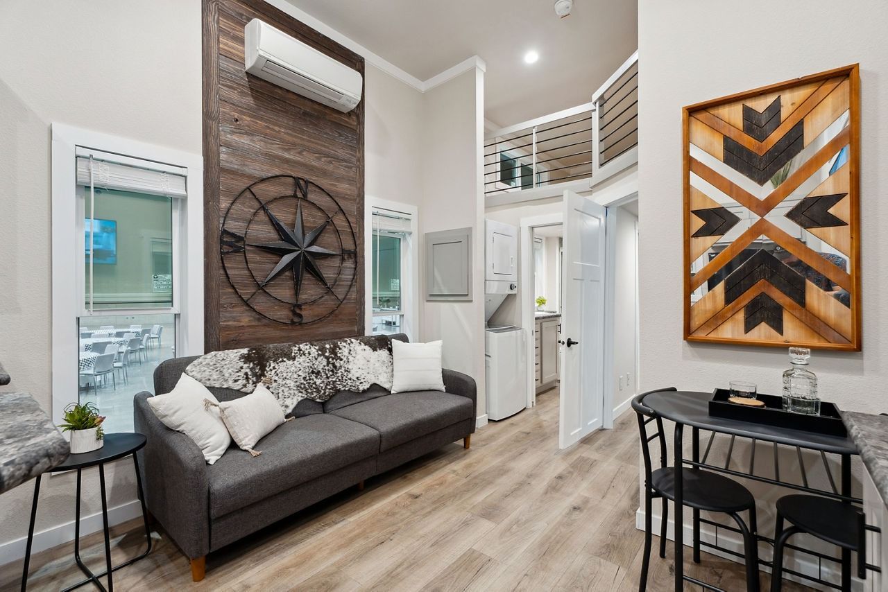 Modern living room with gray sofa, brick accent wall, wall art, and a small dining table by the window.