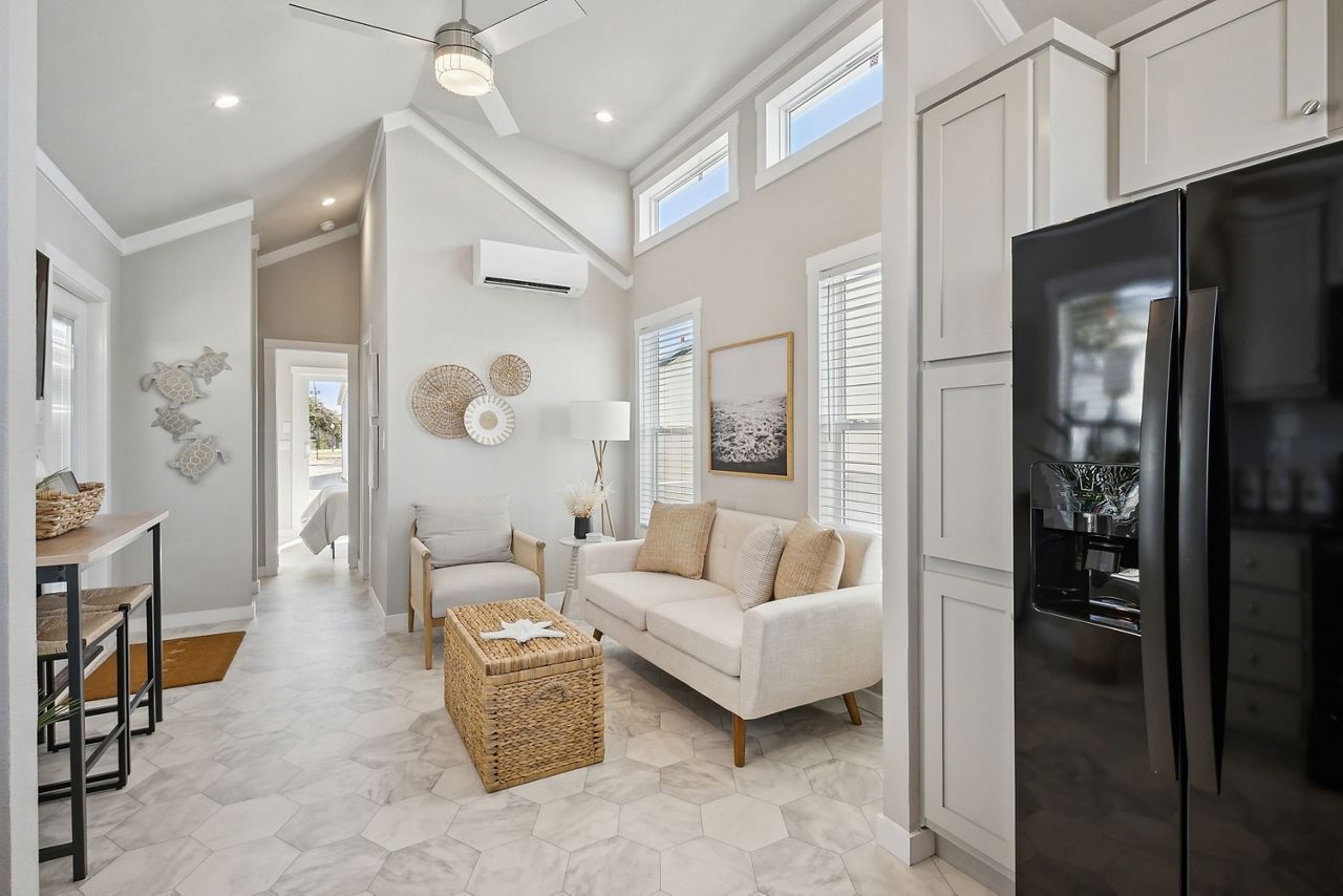 Bright modern living room with high ceiling, white sofa, wicker coffee table, and black refrigerator beside cabinets