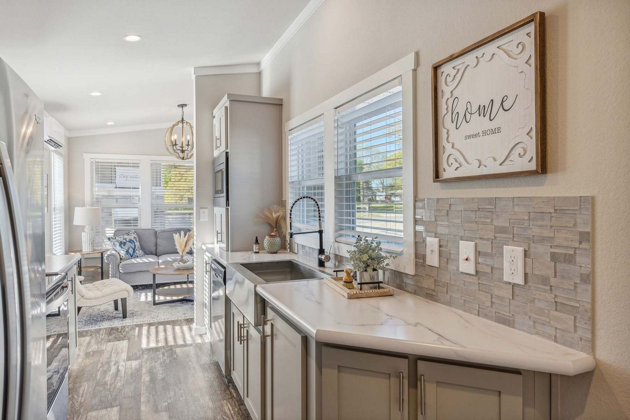 Bright kitchen with gray cabinets, white countertops, tile backsplash, and a view into a sunlit living room.