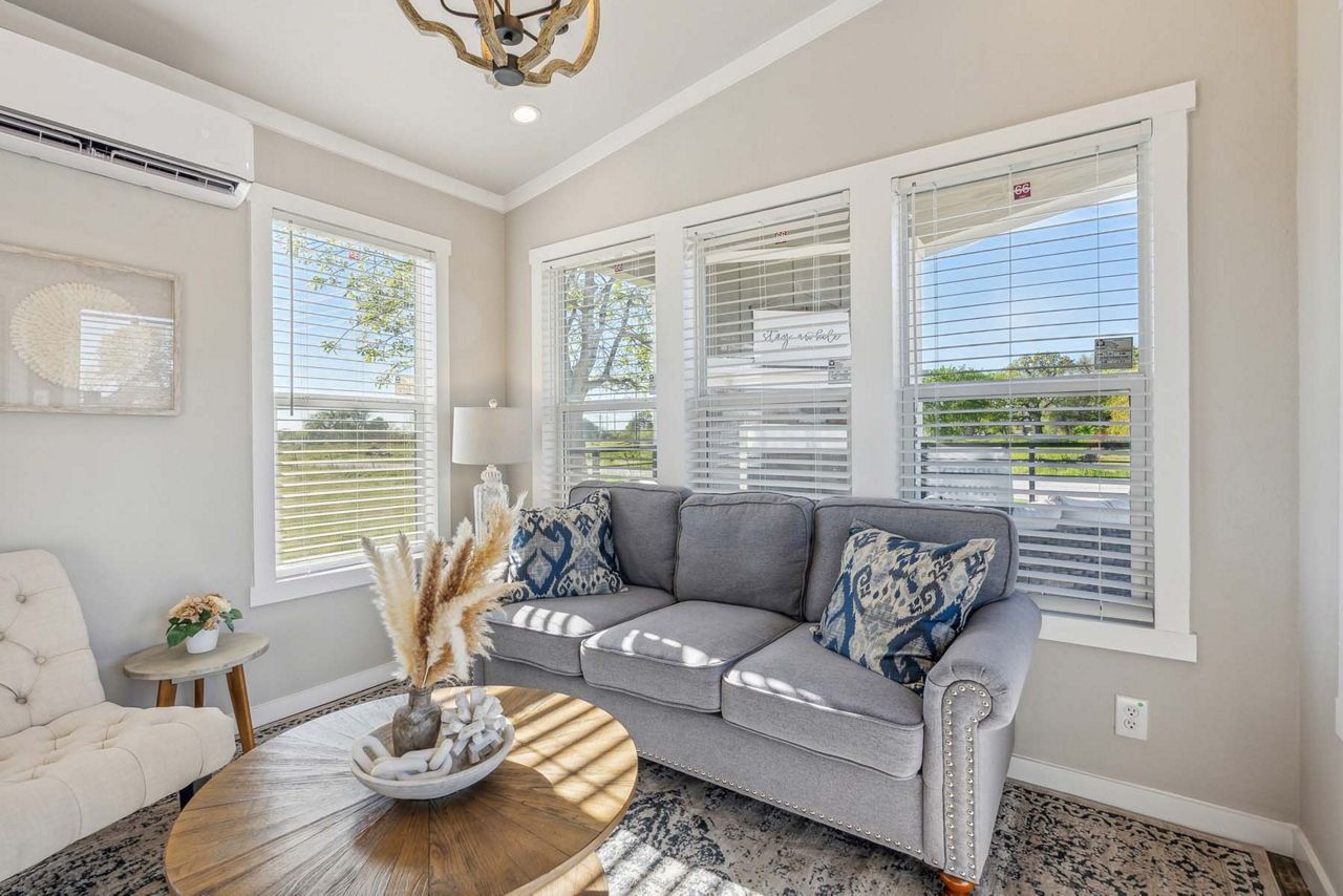 Bright living room with gray sofa, white walls, large windows, and a round coffee table with dried decor.