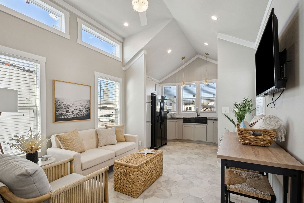 Bright living room with vaulted ceiling, white sofa, woven tables, wall-mounted TV, and open kitchen.